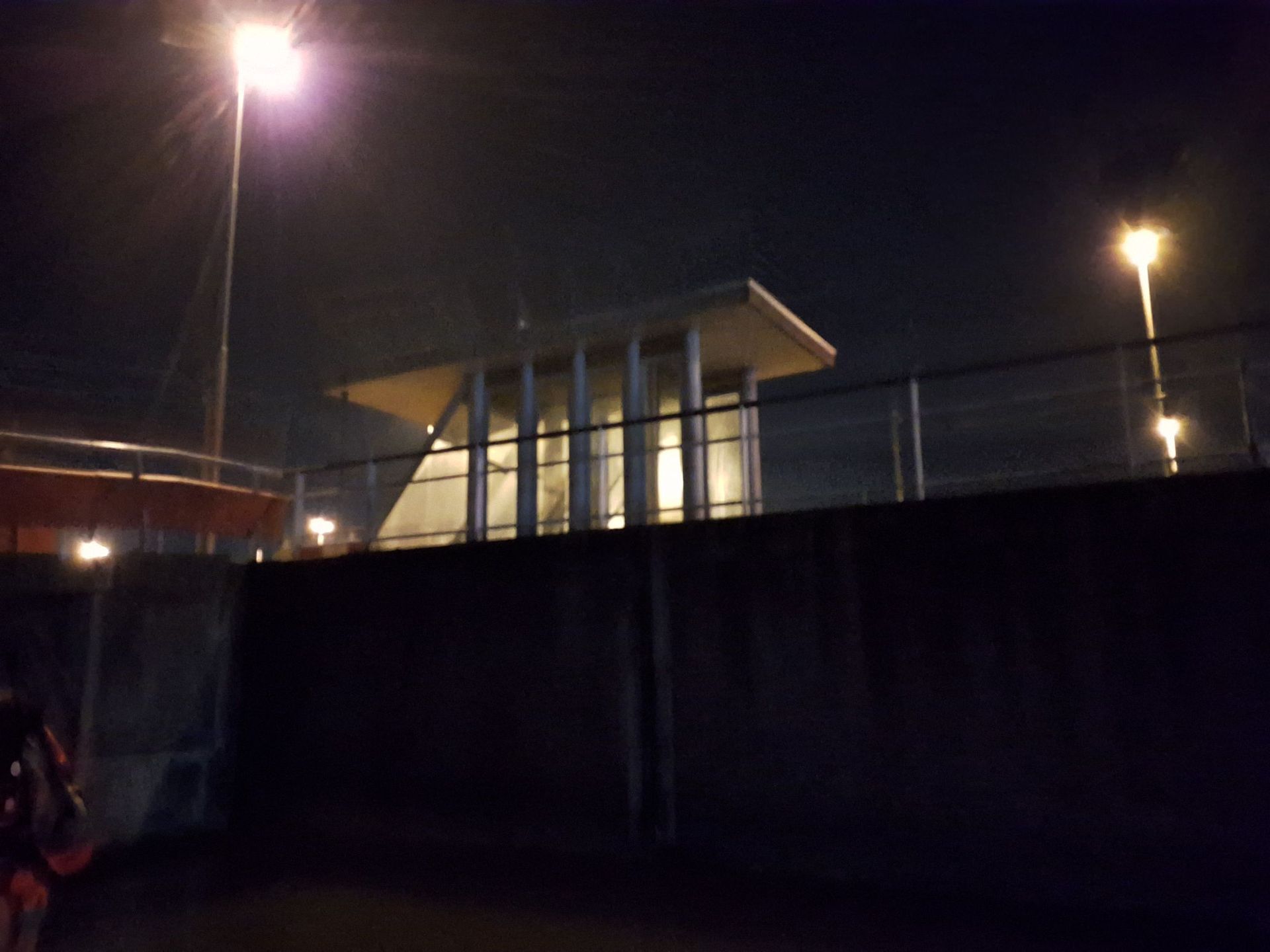 Nachtelijk uitzicht op een verlicht gebouw achter een donkere muur, met straatverlichting erboven.