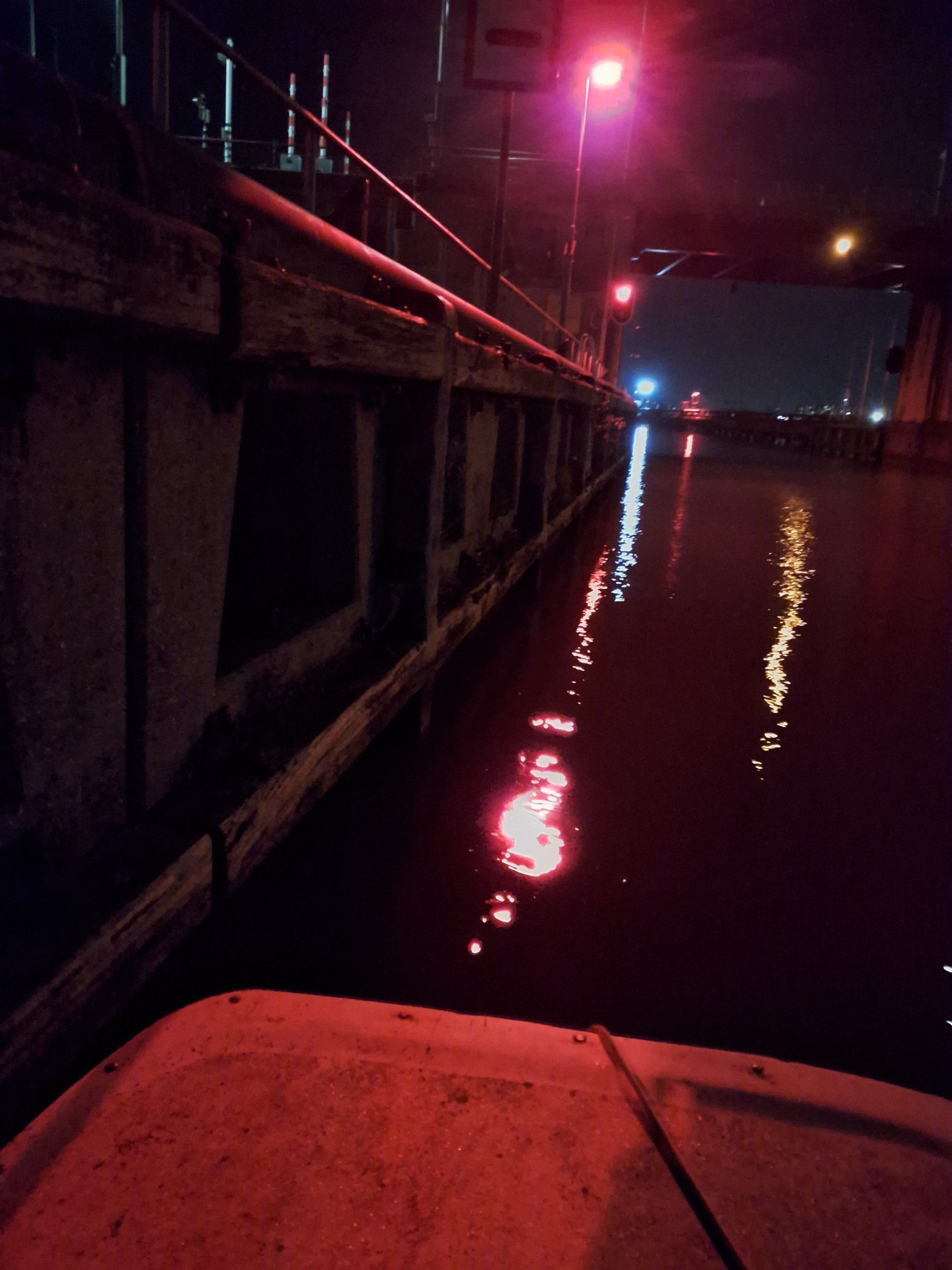 Nachtelijk uitzicht vanaf een boot naast een aanlegsteiger, met rode lichten die reflecteren op het donkere water.