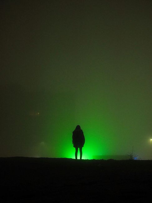 Silhouet van een persoon die op een donkere heuvel staat tegen een groene, mistige gloed in de nacht.