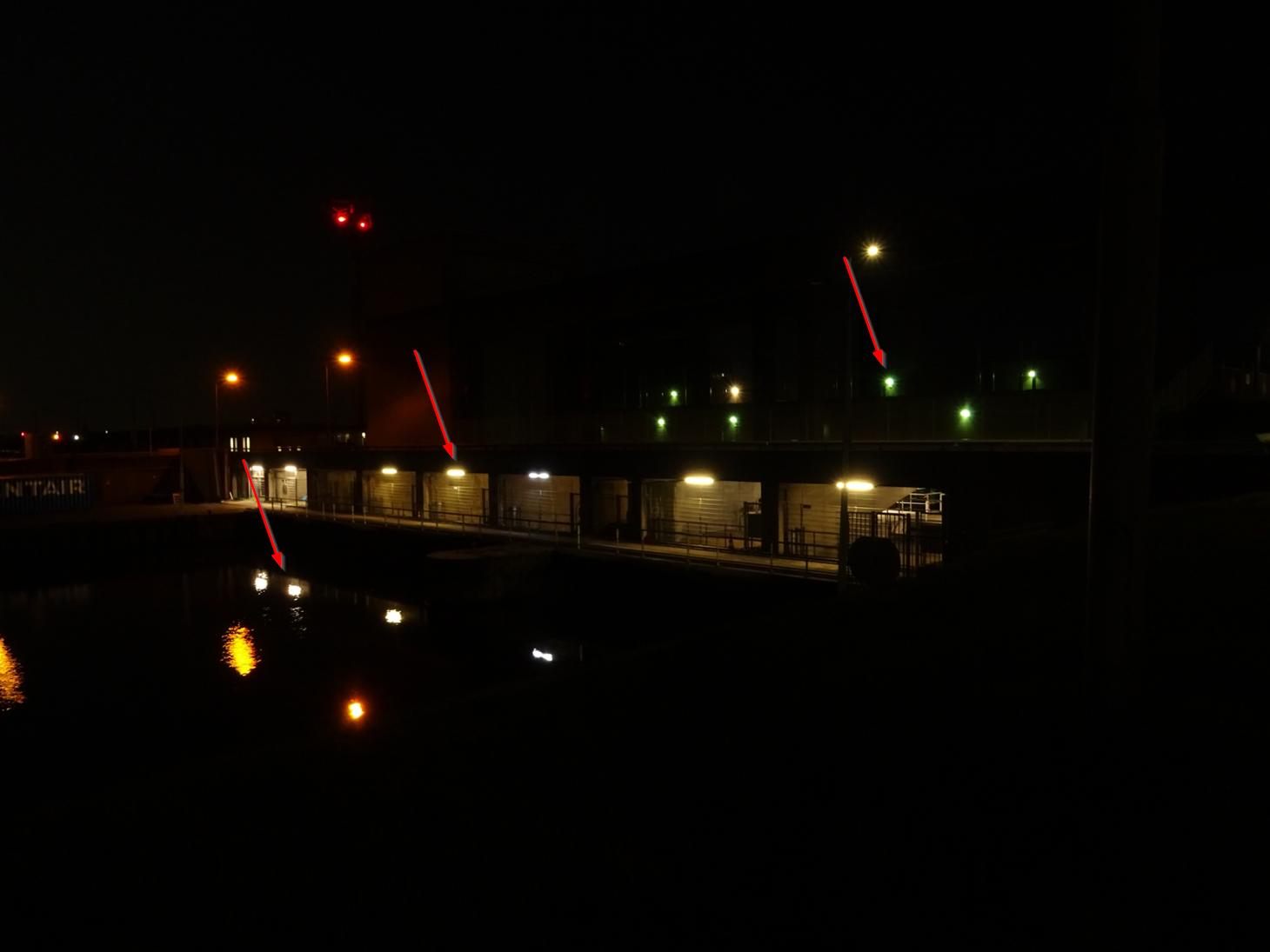 Nachtelijk beeld met verlicht spuigemaal, rode markeringslichten en reflecties op het donkere water.