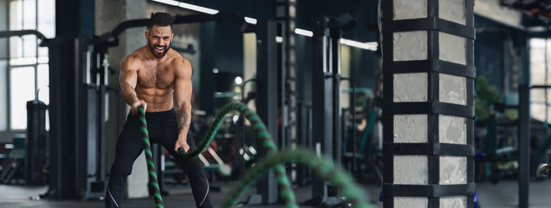 Man working out with battle ropes in a gym; he is shirtless and muscular.