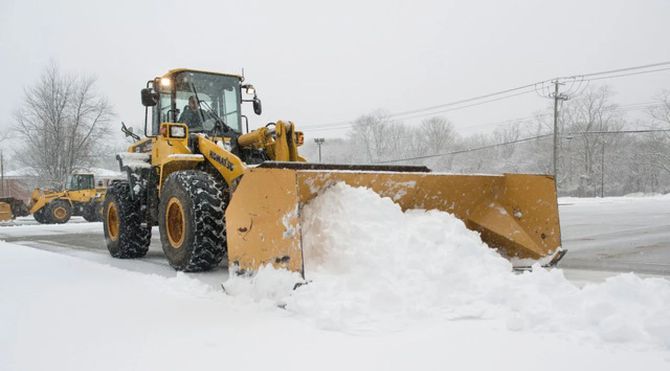 Yellow snowplow clearing deep snow on a foggy road beside bare trees and power lines