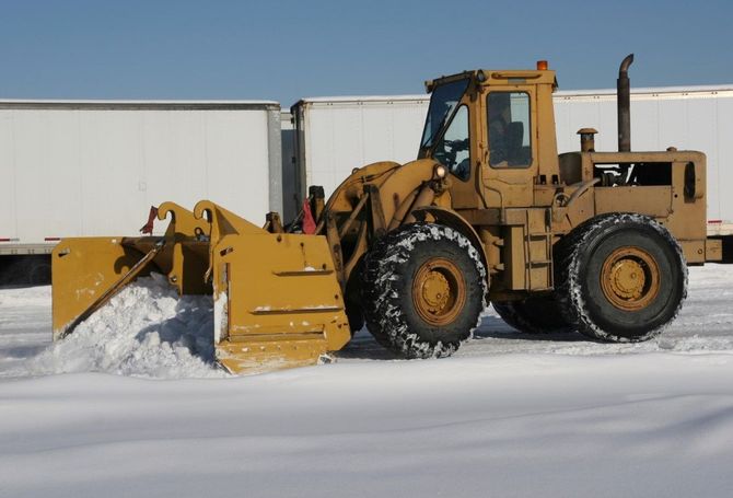 Yellow front-end loader pushing snow in a snowy lot beside white trailers