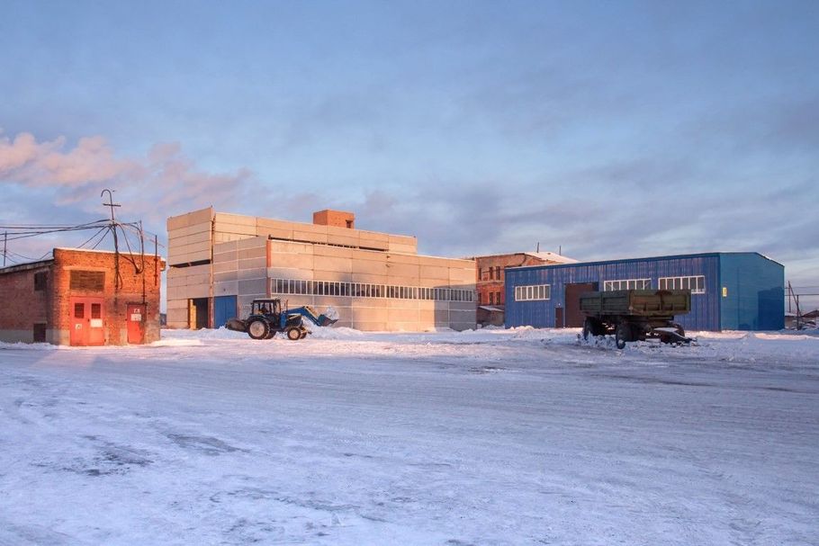 Snowy industrial yard at sunrise with buildings, trucks, and a blue warehouse under a pink sky