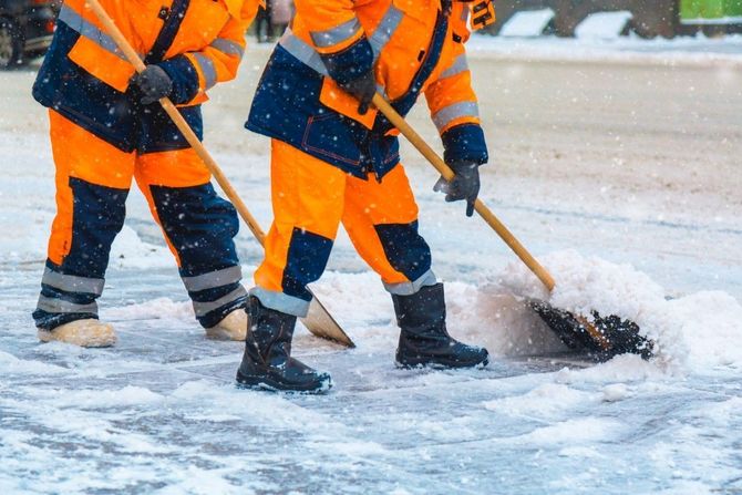 Workers in orange winter gear shoveling snow and ice from a road
