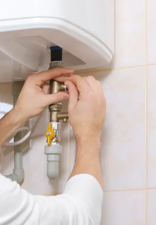 Person adjusting a water heater's valve, inside a tiled room. Hands are on the brass valve, white clothing.