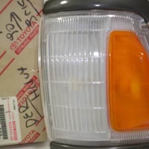 Conjunto de faro delantero de automóvil Toyota con lente transparente y esquina ámbar, junto a una caja de piezas con escritura japonesa.