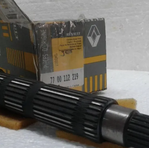 Eje de engranajes de metal negro con una caja de repuestos Renault al fondo sobre una superficie blanca.