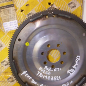Gran volante de inercia de metal negro con borde dentado, marcado en blanco sobre un fondo industrial amarillo.