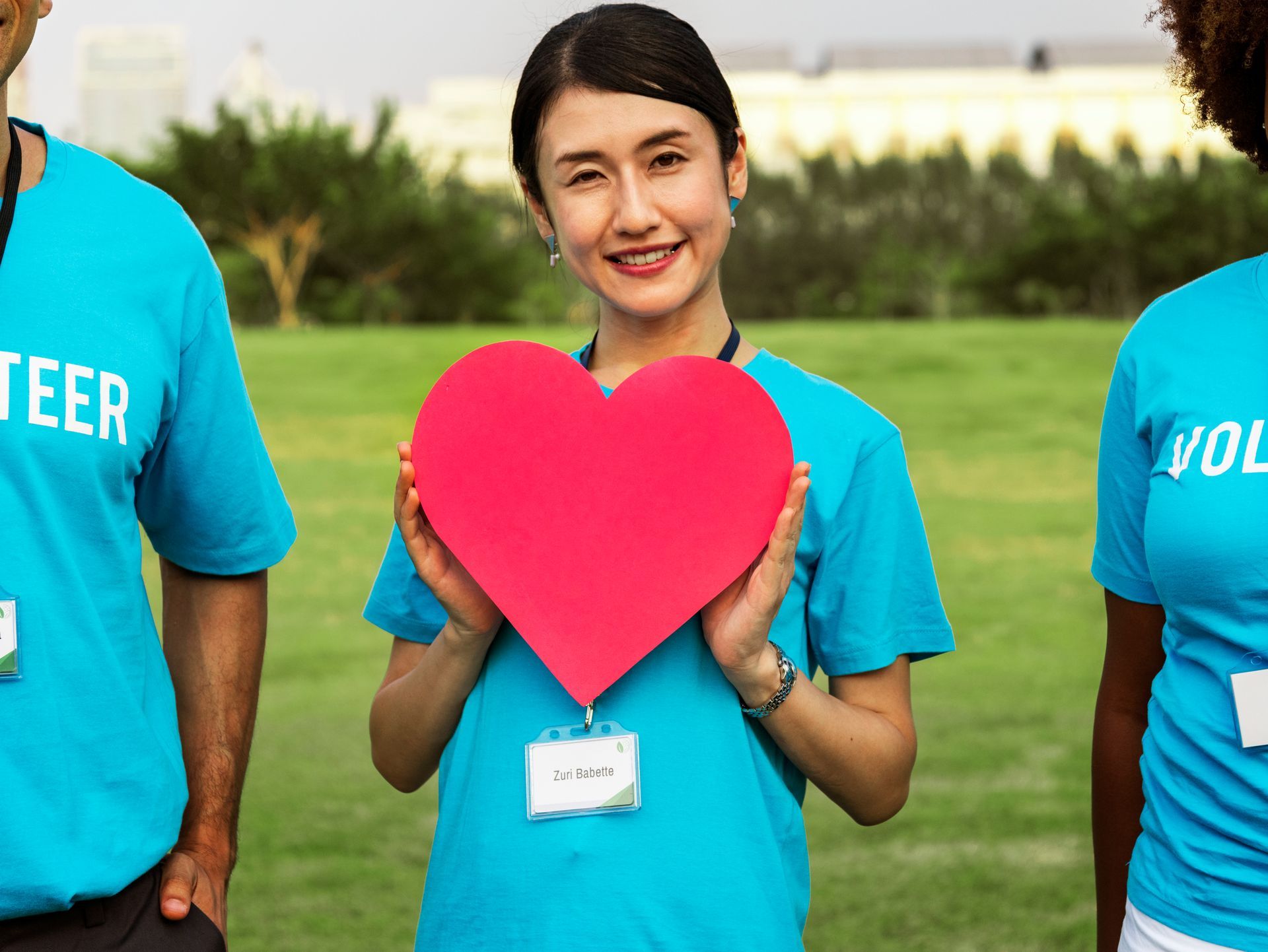 Woman in blue volunteer shirt holds a red heart, smiling outdoors with other volunteers.