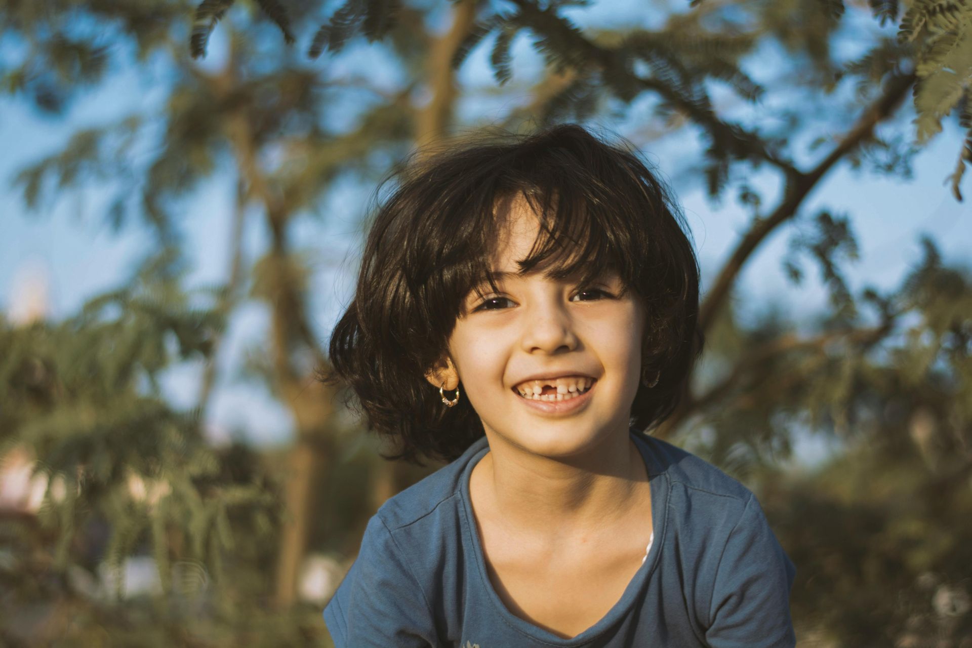 Smiling child with dark hair, missing a front tooth, in front of a tree.