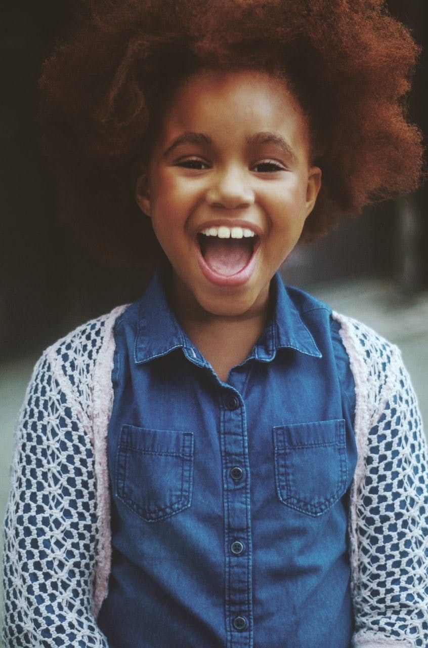 Girl with curly brown hair, smiling widely, wearing a denim shirt and pink cardigan.
