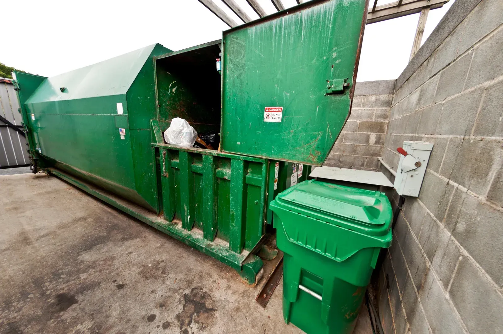 A large green industrial waste compactor with an open door sits next to a green plastic trash bin by a cinder block wall.