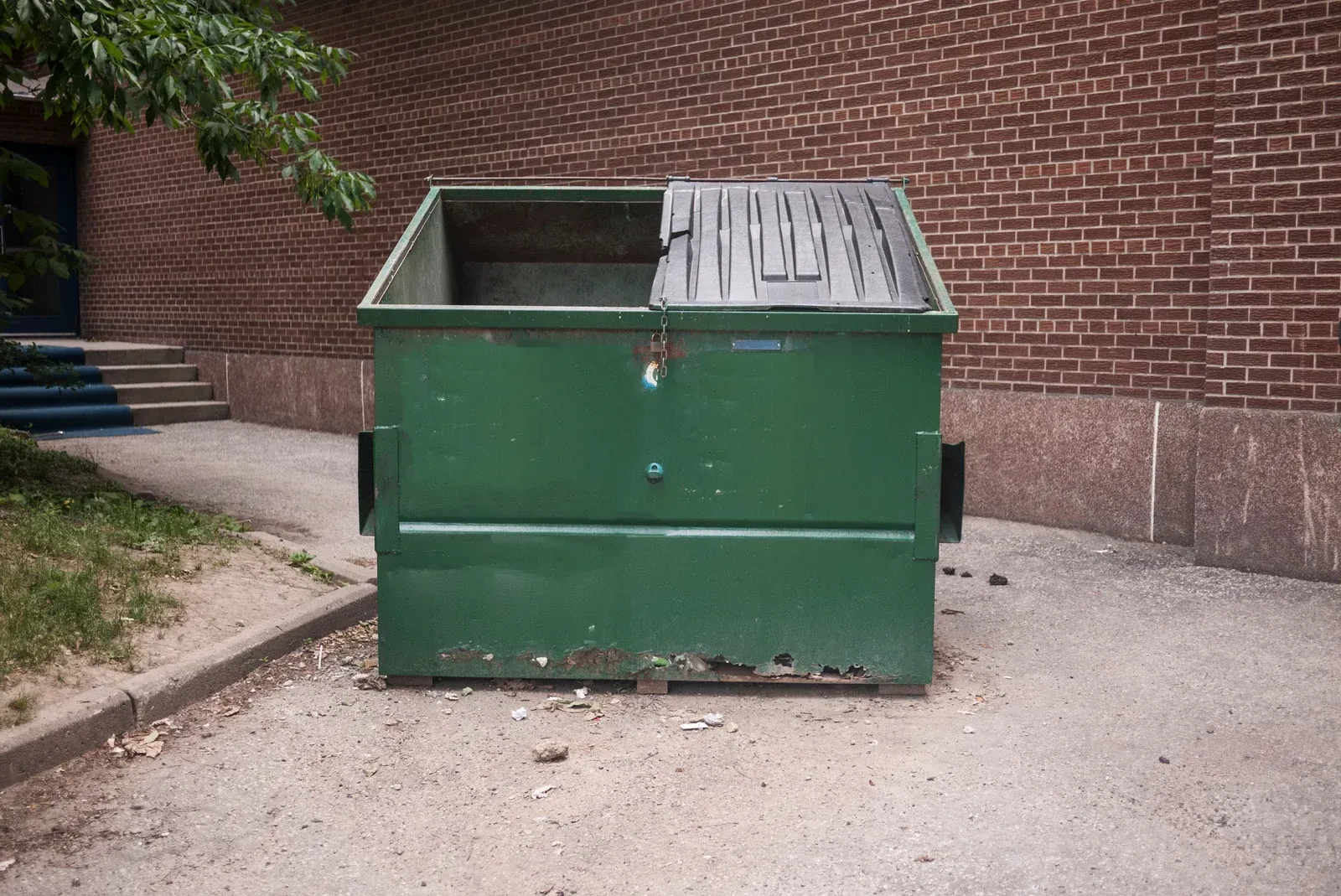 A green metal dumpster with one lid open, sitting on a paved surface against a brick wall.