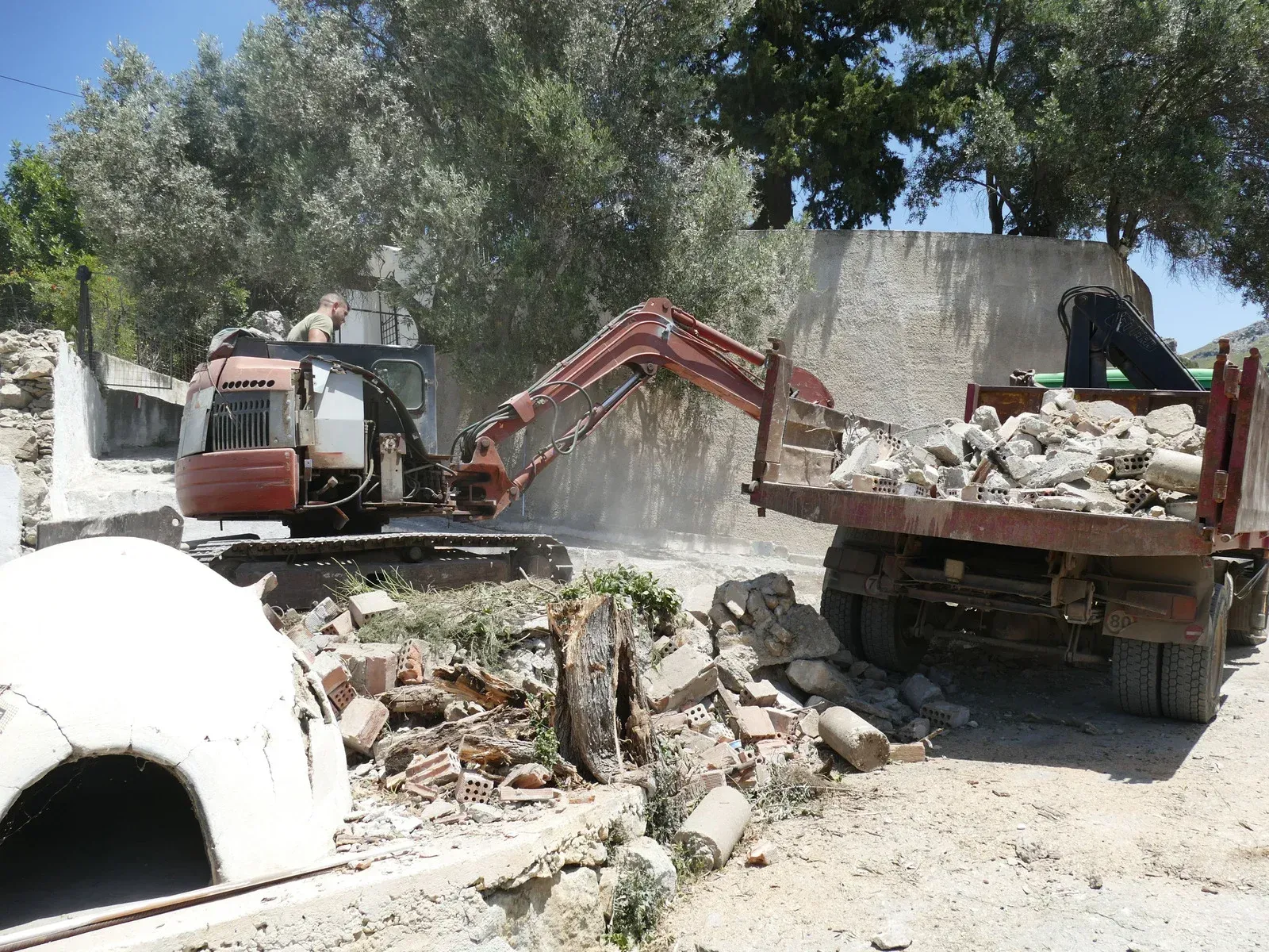 An orange excavator loads debris into the back of a dump truck on a dirt road in front of a stone wall.