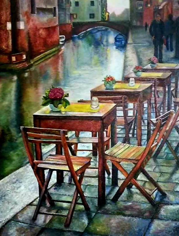 Cafe tables and chairs on a canal-side, flowers, brick buildings, bridge in background, figures walking on a walkway.