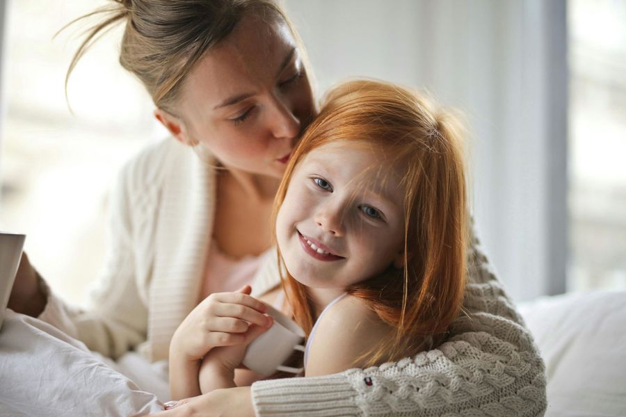 Woman kissing a smiling child with red hair. Both are indoors, possibly on a bed.