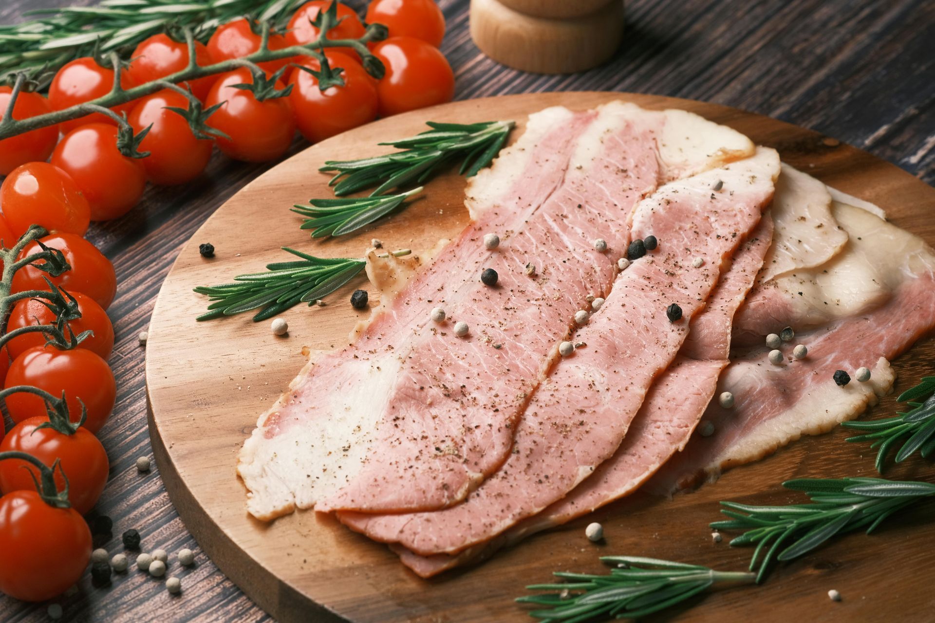 Bacon slices with black pepper on wooden board, sprigs of rosemary, and cherry tomatoes.