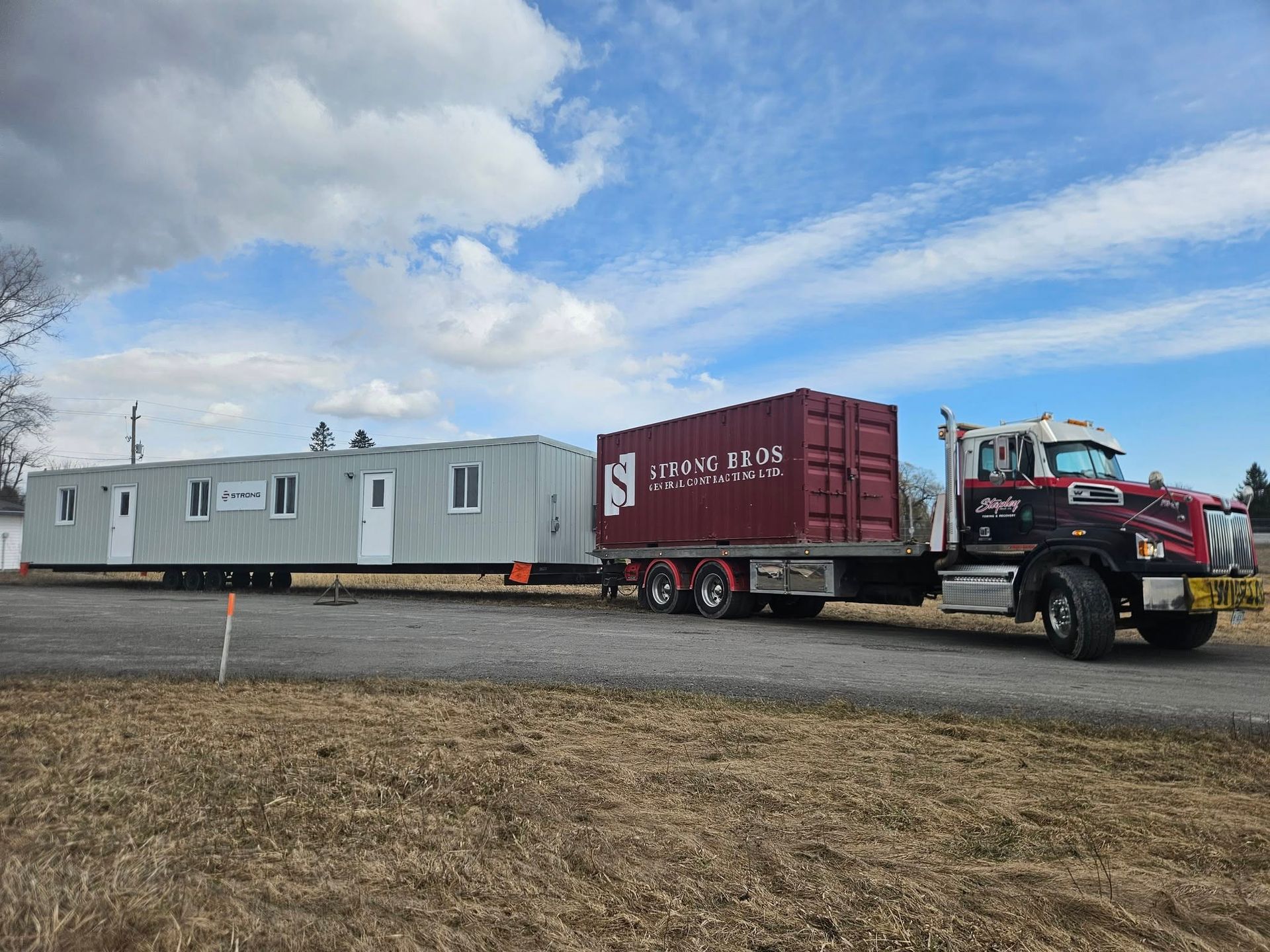Truck transporting a long, modular building and a cargo container on a road, under a cloudy sky.