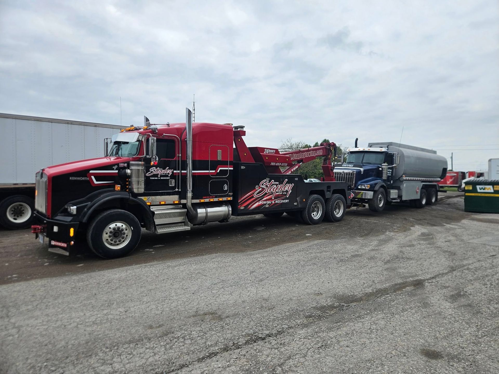 Black and red tow truck towing a tanker truck on a gravel lot under a cloudy sky.