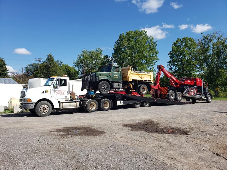 Flatbed truck hauling a green dump truck and red machinery on a sunny day.