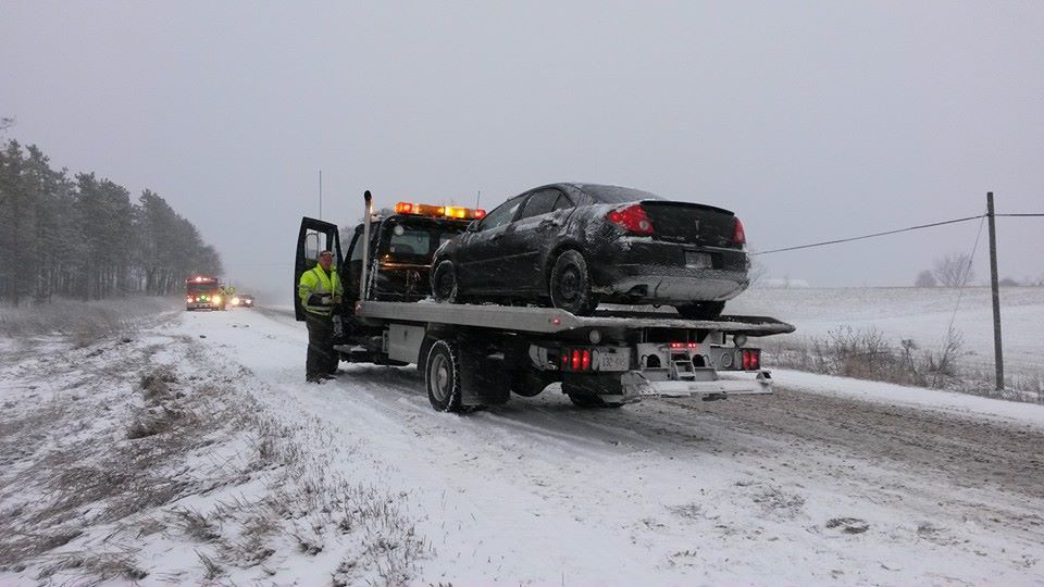 A tow truck loading a black car on a snowy road under a gray sky.