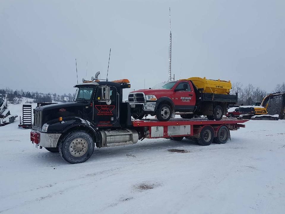 Black tow truck carrying a red truck with a yellow salt spreader in a snowy landscape.