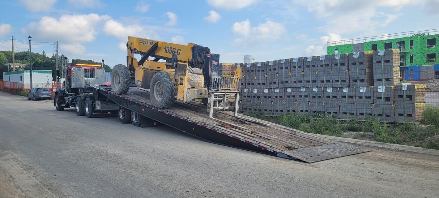 Truck transporting a yellow construction vehicle on a flatbed, alongside stacked gray blocks.