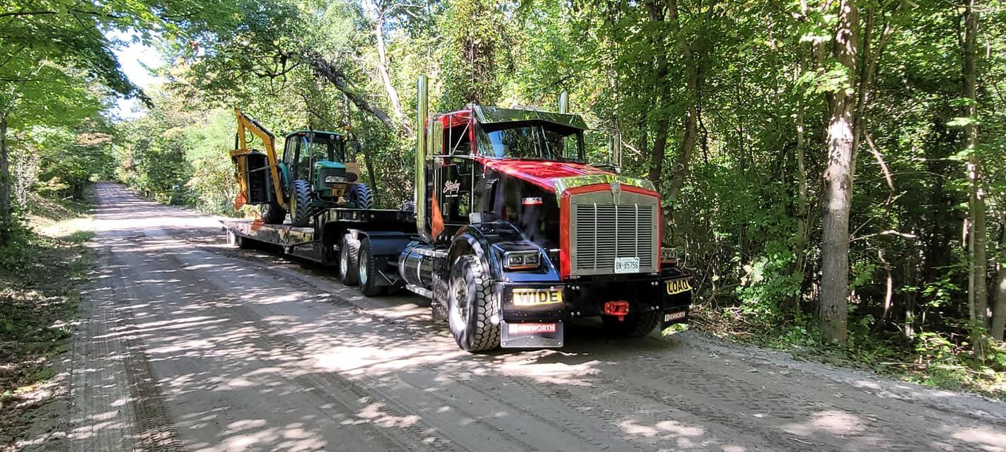 A large red semi-truck hauling a yellow excavator on a gravel road surrounded by trees.
