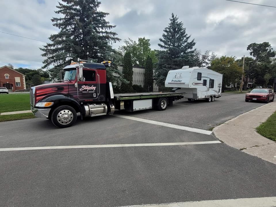 Tow truck hauling a white camper on a residential street. Truck is black and red. A car is on the right.