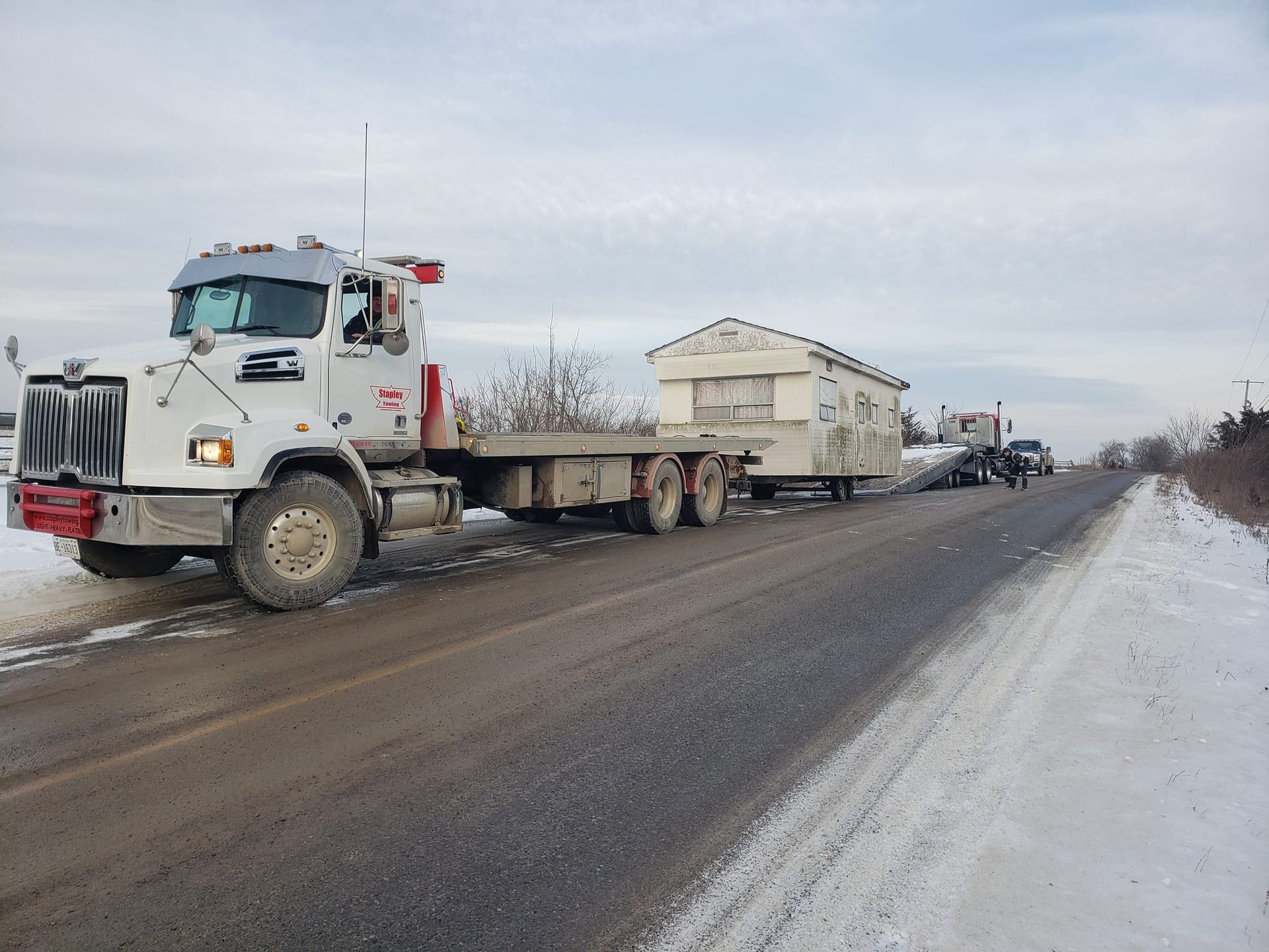 White semi-truck transporting a small building on a flatbed trailer along a snowy road.