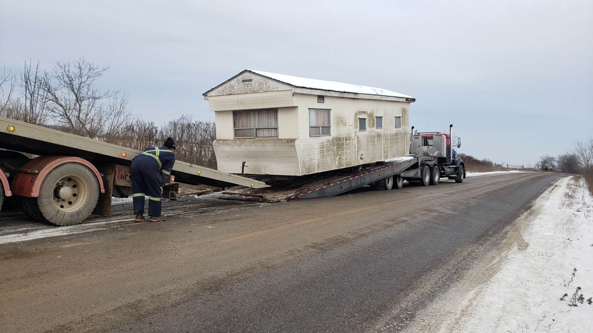 Mobile home being transported on a flatbed trailer on a snowy road, two people assisting.