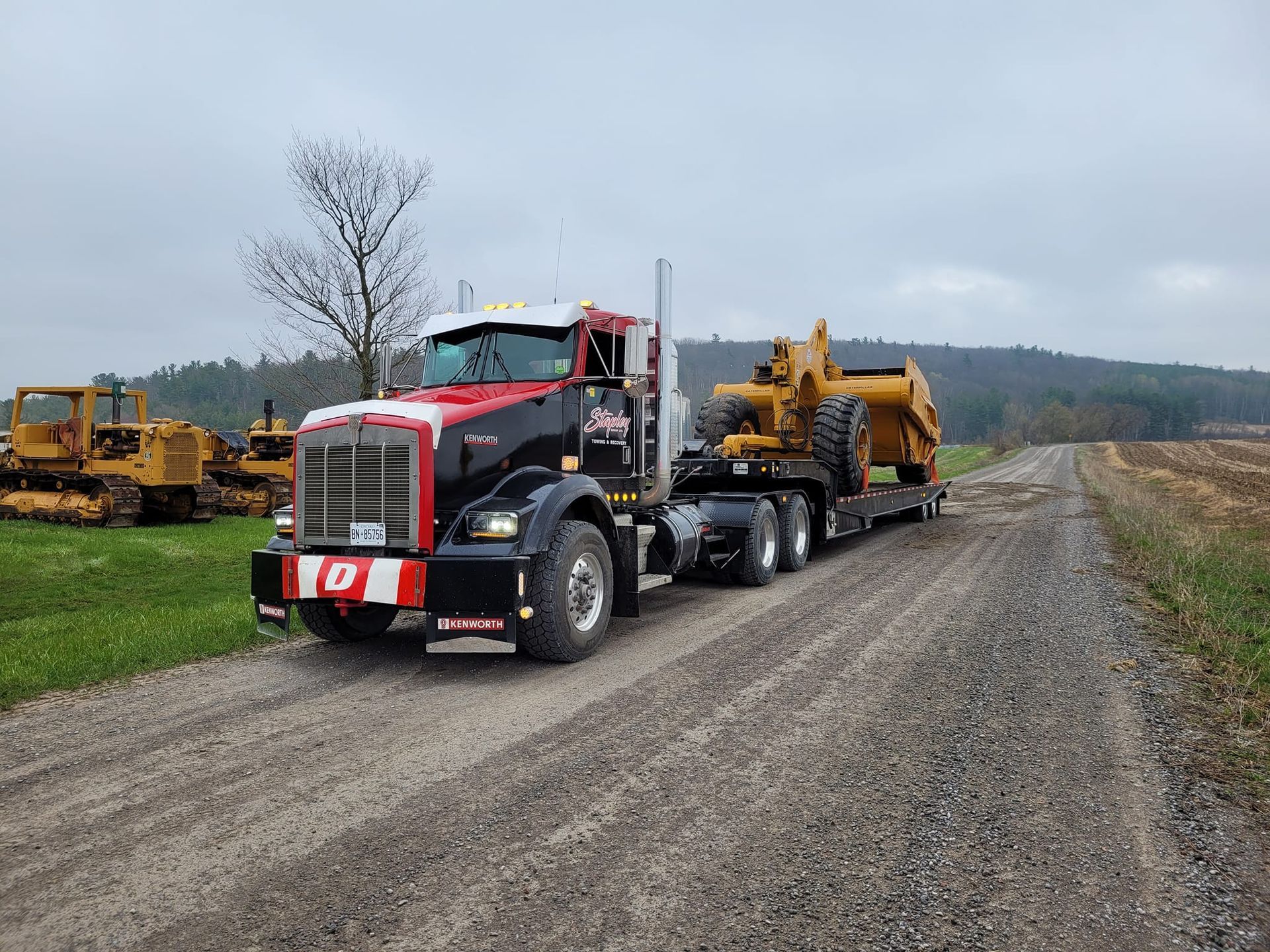 Red and black semi-truck hauling a yellow piece of heavy machinery down a gravel road.