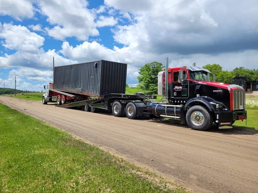 Black semi-truck hauling large, black container on dirt road under blue sky.