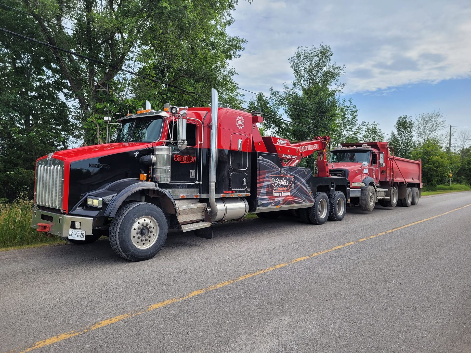A red and black tow truck pulling a red dump truck on a road near trees.