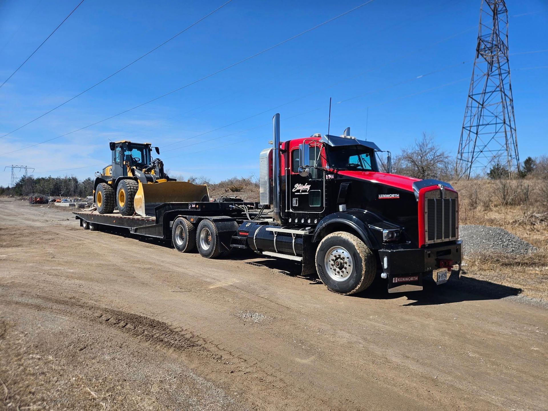 Black and red semi-truck hauling a yellow construction loader on a lowboy trailer on a dirt road.