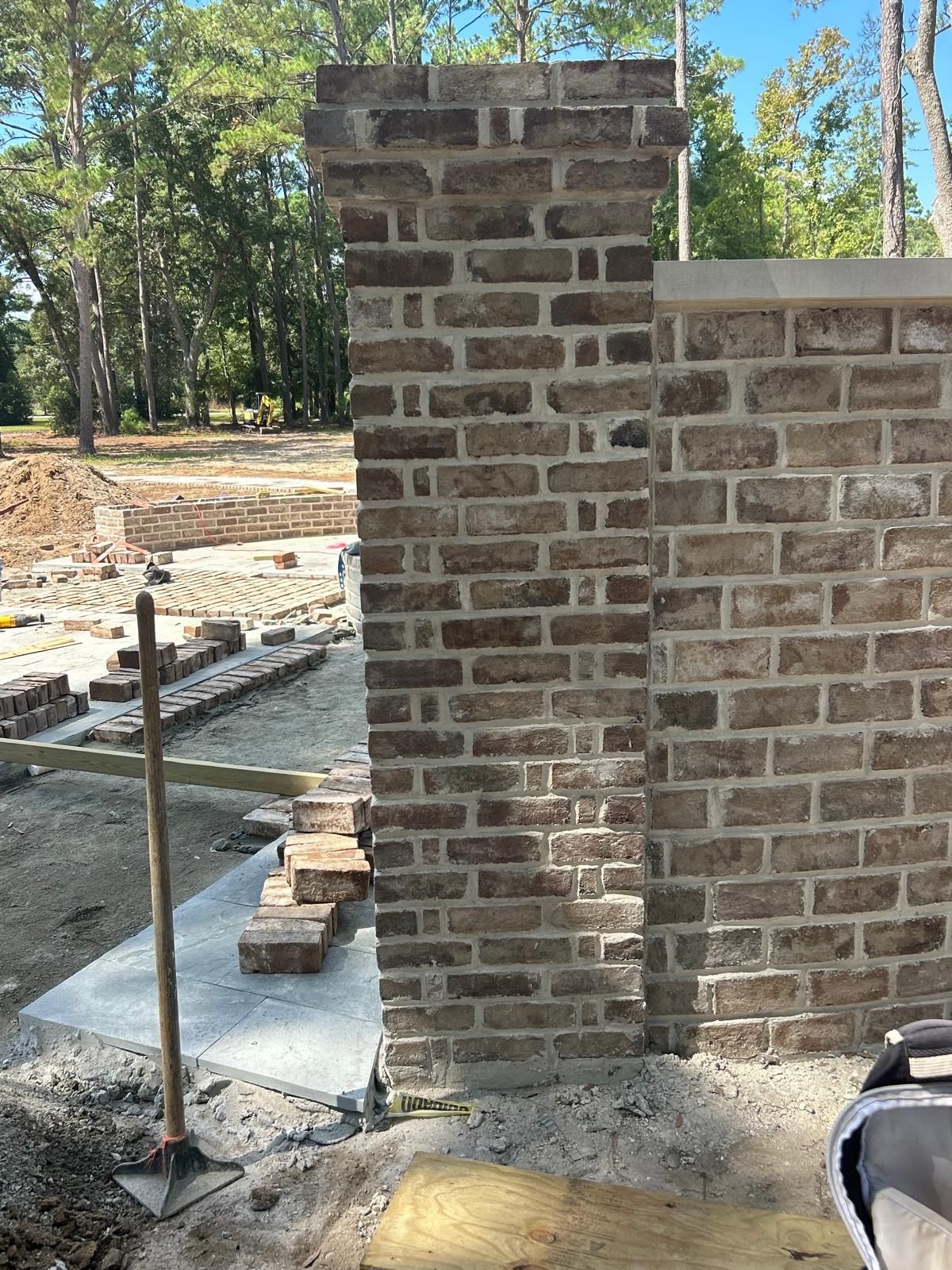 Brick pillar and wall under construction outdoors. Background of trees and dirt.