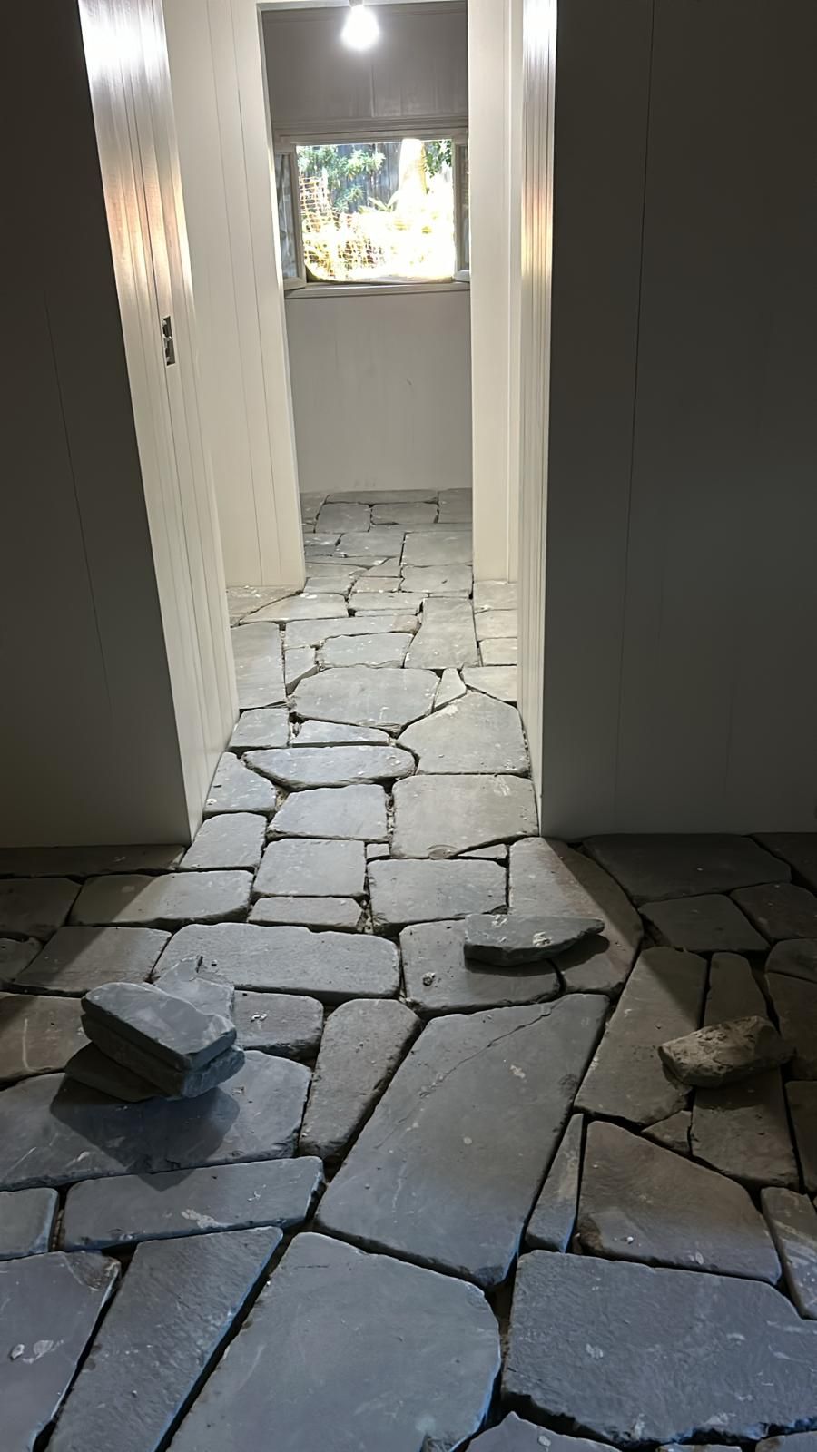 Hallway with dark gray, irregular stone tile flooring; light gray walls; natural light from window at end.