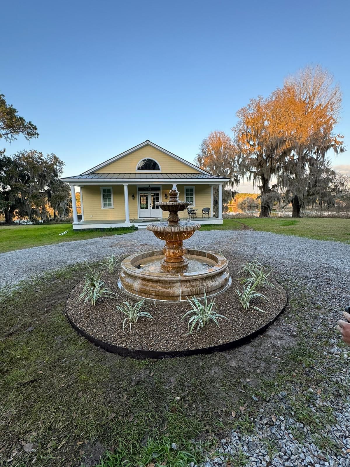 Yellow house with a fountain in front, on a gravel driveway, against a grassy yard under a blue sky.