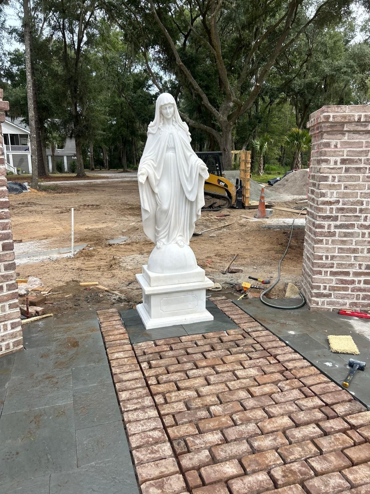 Statue of Virgin Mary on a brick path, between brick pillars. Construction tools visible.