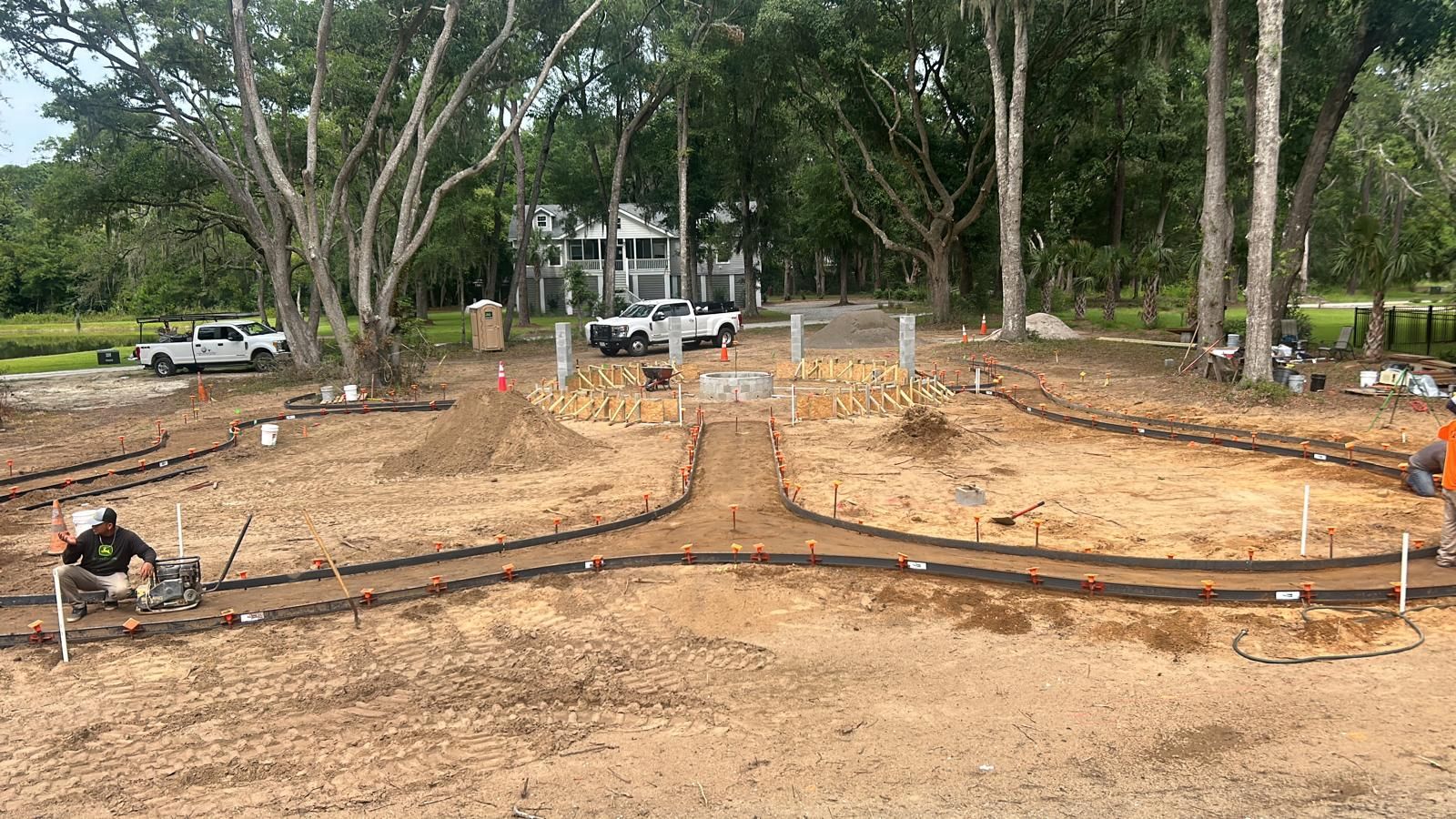Construction site with workers, trucks, and materials. Paths are being laid in a dirt yard with trees and a house in the background.