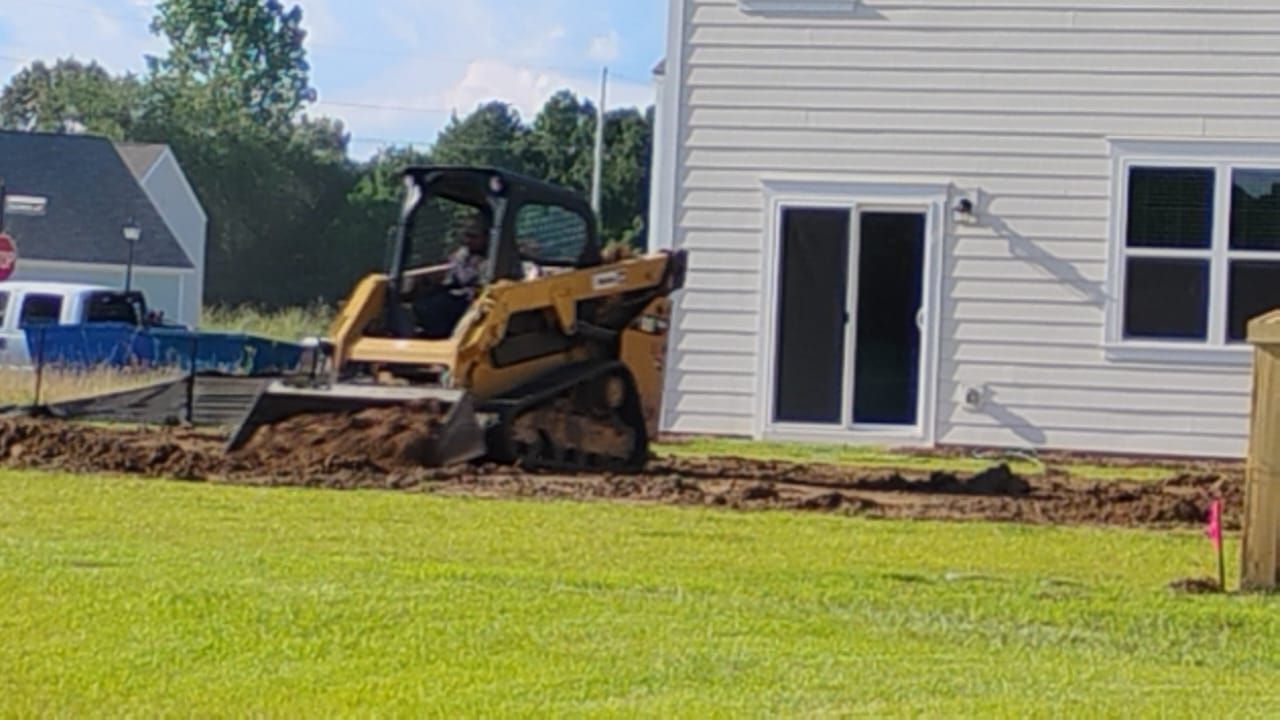Yellow skid steer digging trench near a white house with sliding glass doors on a sunny day.