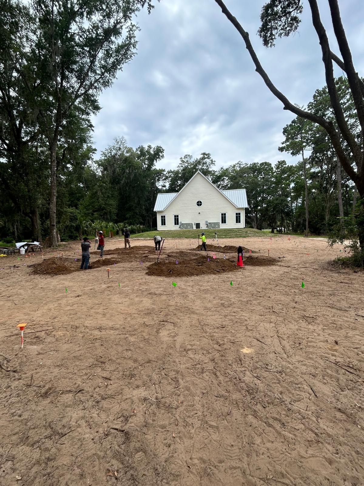 People working on landscaping in front of a white building with a peaked roof, surrounded by trees.