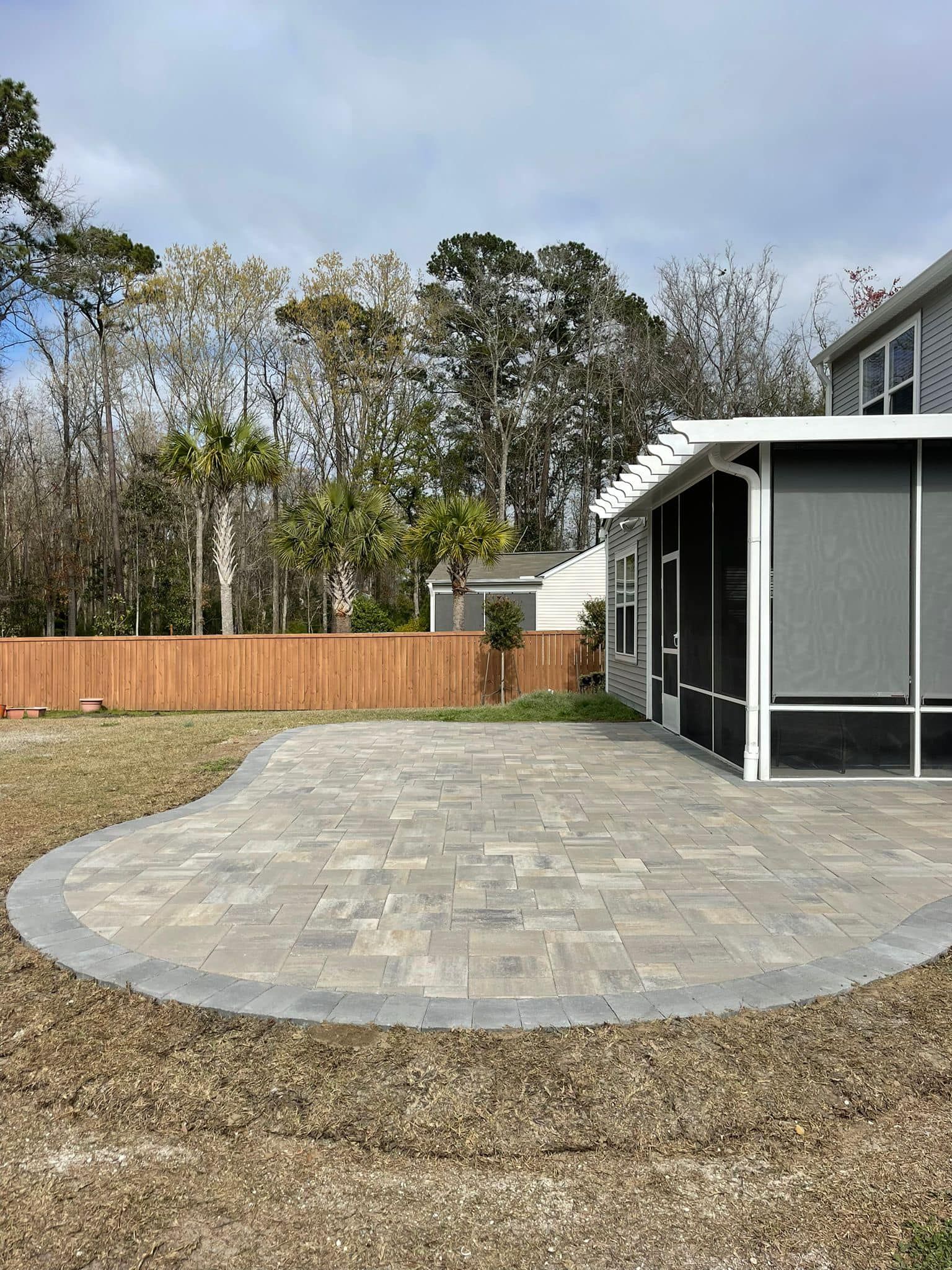Patio with pavers and dark border, screened-in porch, wooden fence, trees, and cloudy sky.