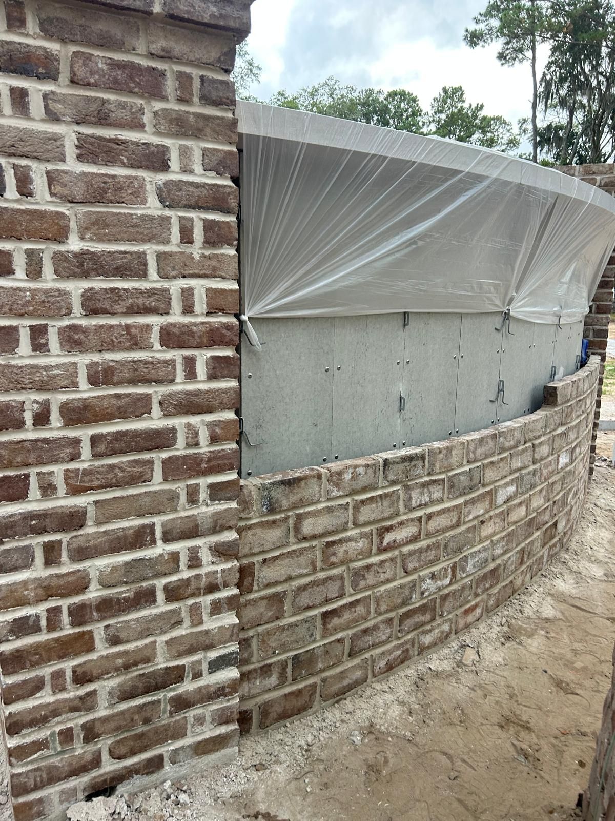 Brick wall under construction, partially curved. Brown bricks, gray mortar. Plastic sheeting covers unfinished section.