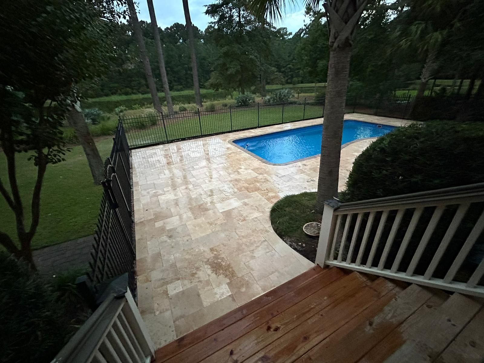 Pool area: blue pool, tan stone patio, wooden stairs, black fence, green grass and trees.