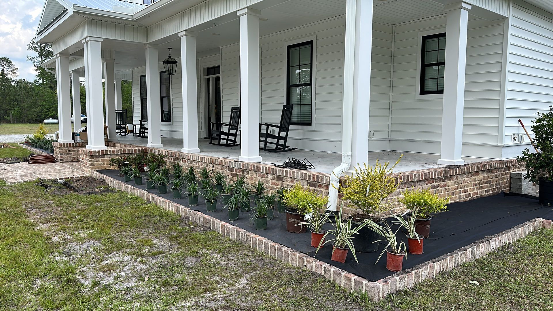 White house with a wrap-around porch, brick base, and a flower bed filled with potted plants.
