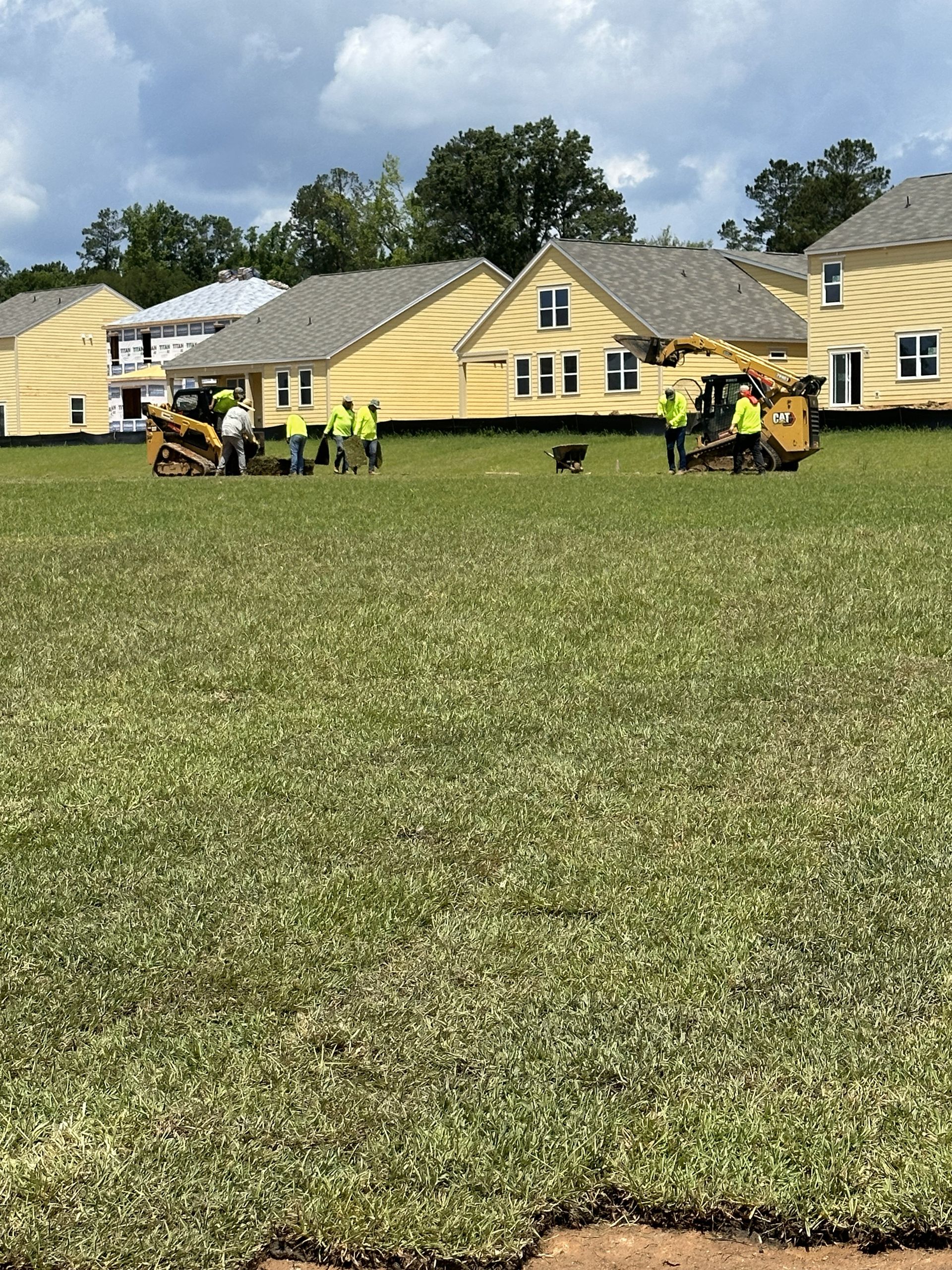Workers in yellow vests operate machinery on a grassy field in front of yellow houses.