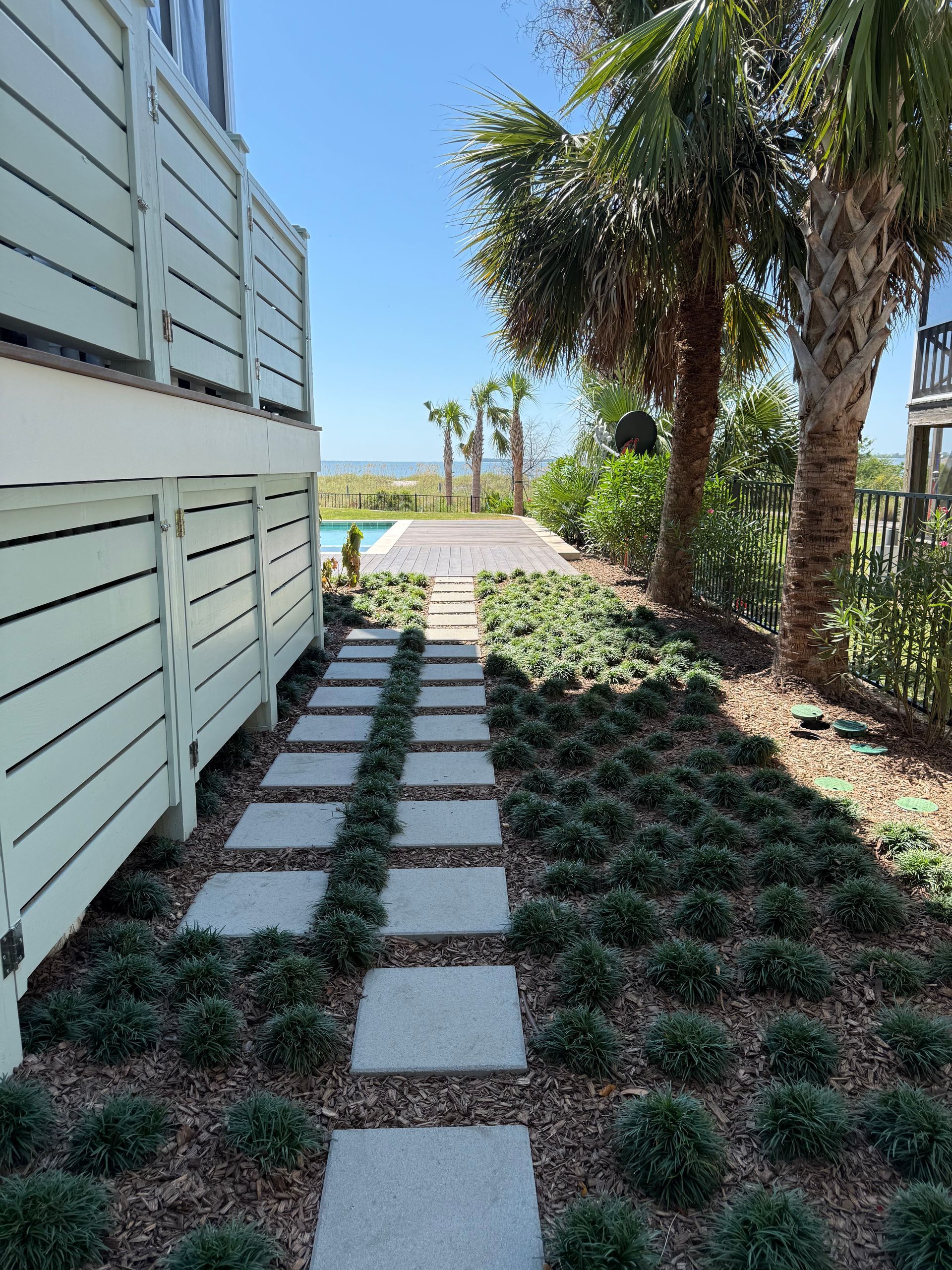 Stone path through landscaped yard leading to the ocean. White fence on the left and palm trees on the right.