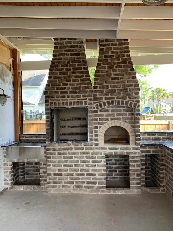 Outdoor brick fireplace with oven and sink under a white-beamed ceiling.